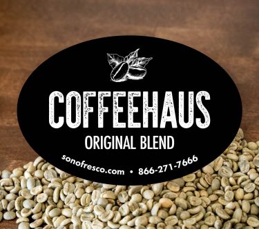 Black oval label reading ‘COFFEEHAUS Original Blend’ with a coffee bean illustration. Background shows green coffee beans on a wooden surface.