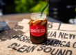 A glass of iced coffee on a green coffee burlap bag under the sunlight, condensation forming on the glass, ice cubes melting, creating a refreshing scene.