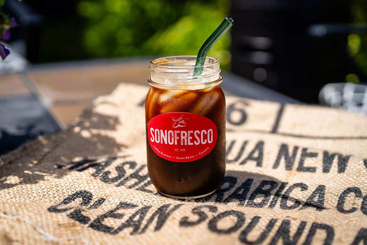 A glass of iced coffee on a green coffee burlap bag under the sunlight, condensation forming on the glass, ice cubes melting, creating a refreshing scene.