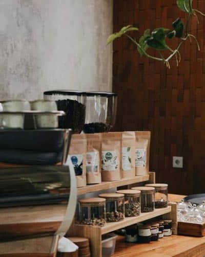 Coffee station with jars of spices, pouches of roasted beans, and a grinder setup on a wooden counter in a cozy café space.